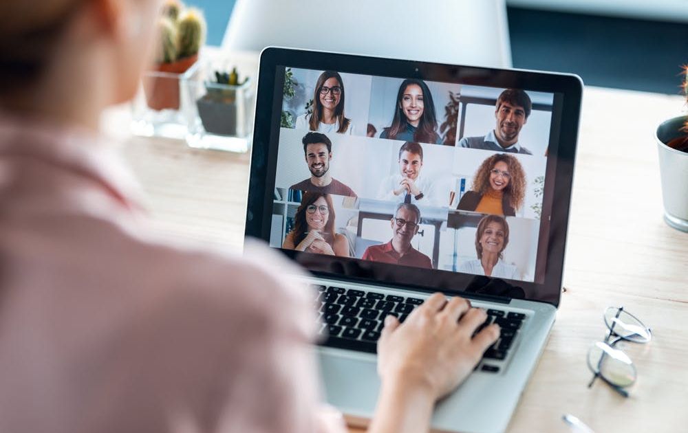 Een over-de-schouder-weergave van een vrouw die deelneemt aan een videovergadering op haar laptop, waarop een raster van negen glimlachende, diverse collega's te zien is.