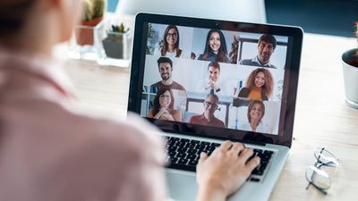 Een over-de-schouder-weergave van een vrouw die deelneemt aan een videovergadering op haar laptop, waarop een raster van negen glimlachende, diverse collega's te zien is.