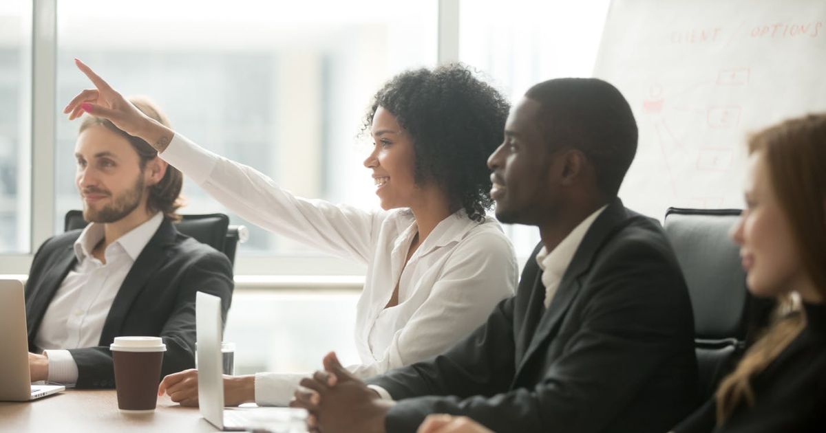 Een lachende Zwarte vrouw die haar hand opsteekt om een vraag te stellen of een bijdrage te leveren tijdens een zakelijke vergadering, zittend aan een vergadertafel naast diverse collega's.