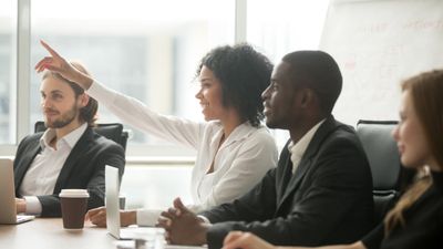 Een lachende Zwarte vrouw die haar hand opsteekt om een vraag te stellen of een bijdrage te leveren tijdens een zakelijke vergadering, zittend aan een vergadertafel naast diverse collega's.