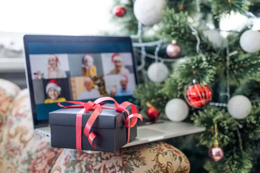 A wrapped gift box sits on a laptop in front of a decorated Christmas tree, while a video call with several people wearing Santa hats appears on the screen, suggesting a virtual holiday celebration.