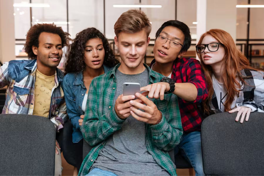 A diverse group of five young friends crowding together in a room, looking curiously at a smartphone held by a young man in the center.