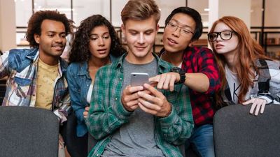 A diverse group of five young friends crowding together in a room, looking curiously at a smartphone held by a young man in the center.