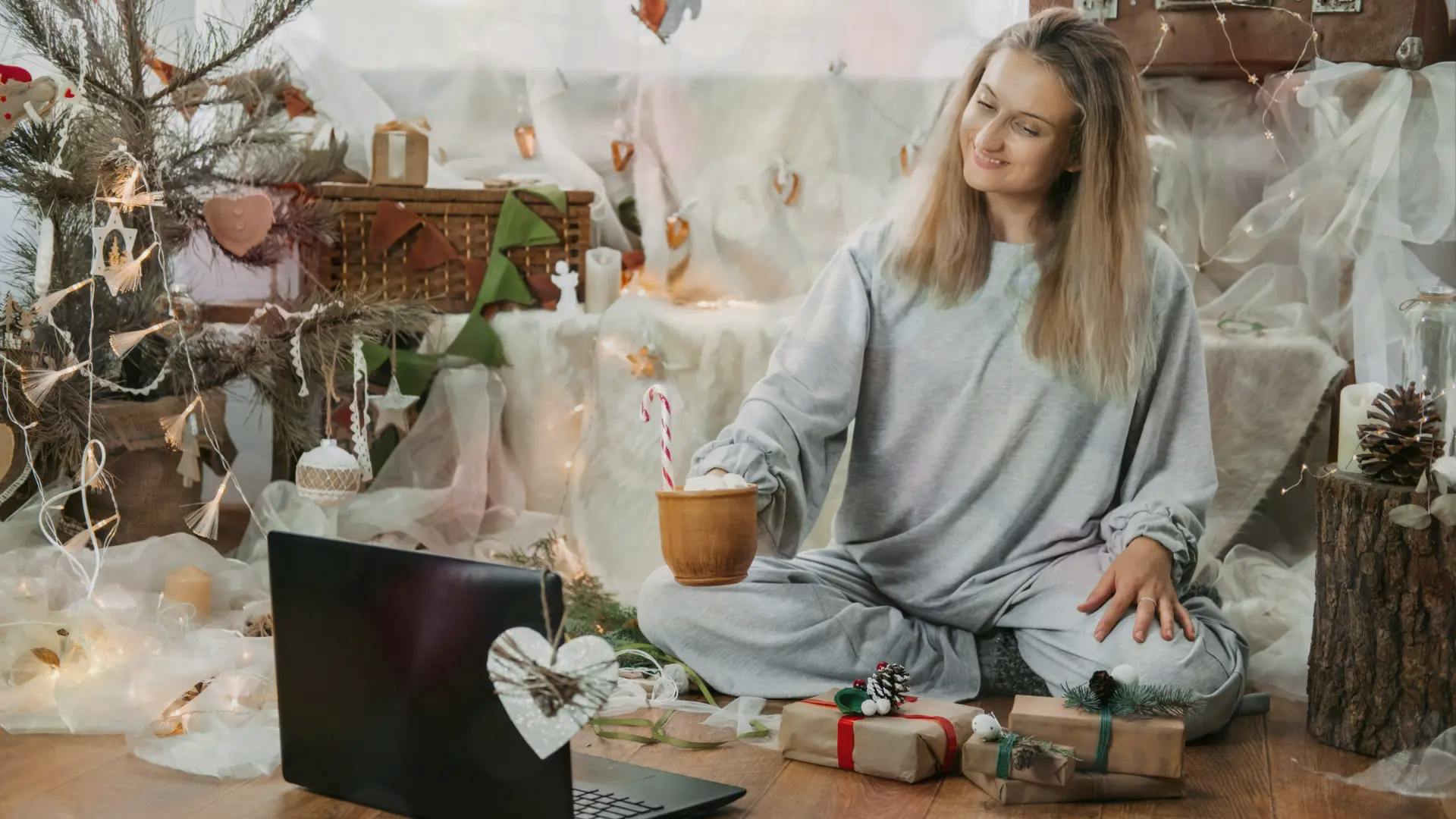 Une femme souriante en pyjama gris assise en tailleur sur le sol, tenant une tasse avec un sucre d'orge tout en regardant un ordinateur portable ouvert dans une piece decoree pour les fetes.