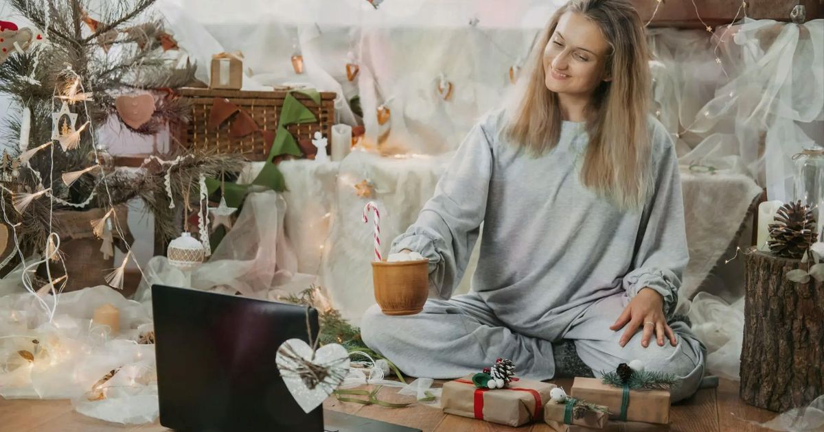 A smiling woman in grey pajamas sitting cross-legged on the floor, holding a mug with a candy cane while looking at an open laptop in a festively decorated holiday room.