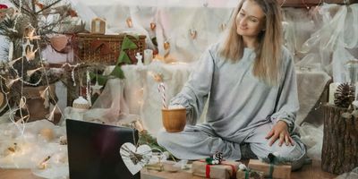 Een glimlachende vrouw in grijze pyjama die met gekruiste benen op de vloer zit, een mok met een zuurstok vasthoudt en naar een open laptop kijkt in een feestelijk versierde kerstomgeving.