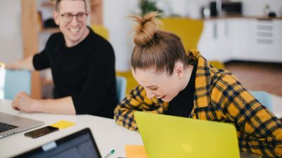 A young woman in a yellow flannel shirt laughs while looking down at her lime green laptop, while a male colleague in a black shirt smiles in the background at an office desk.