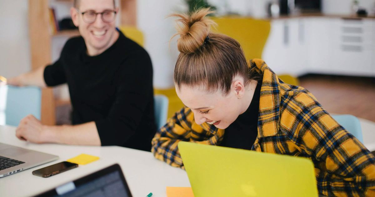 A young woman in a yellow flannel shirt laughs while looking down at her lime green laptop, while a male colleague in a black shirt smiles in the background at an office desk.