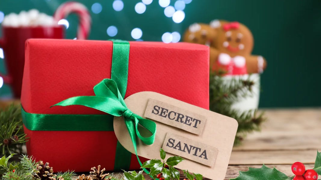 Gros plan d'un cadeau de Noël rouge enrubanné d'un ruban de satin vert et d'une étiquette portant la mention "SECRET SANTA", sur un fond de fête légèrement flou.