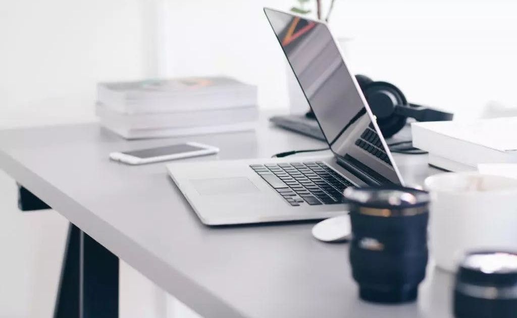 Een moderne, minimalistische werkplek met een open laptop, een smartphone, een stapel boeken en een koptelefoon op een strakke grijze bureau.