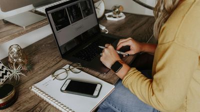 A woman in a yellow sweater working on a laptop at a rustic wooden desk, with an open notebook, smartphone, and glasses resting nearby.