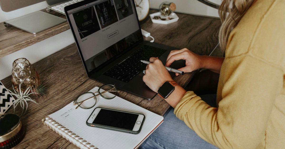 A woman in a yellow sweater working on a laptop at a rustic wooden desk, with an open notebook, smartphone, and glasses resting nearby.