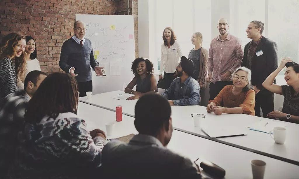 A large, diverse group of smiling people gathered around a long conference table for a collaborative business workshop or team meeting.