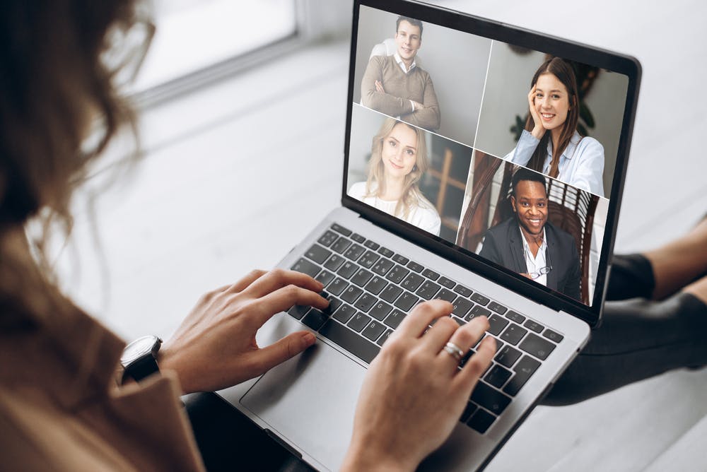 Een over-de-schouder weergave van een vrouw die deelneemt aan een videoconferentiegesprek op haar laptop, met vier glimlachende, diverse collega's op het scherm.