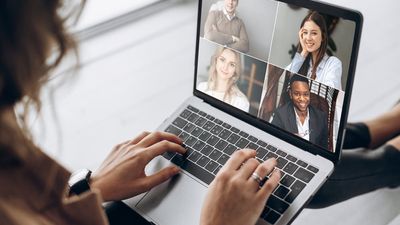 Een over-de-schouder weergave van een vrouw die deelneemt aan een videoconferentiegesprek op haar laptop, met vier glimlachende, diverse collega's op het scherm.