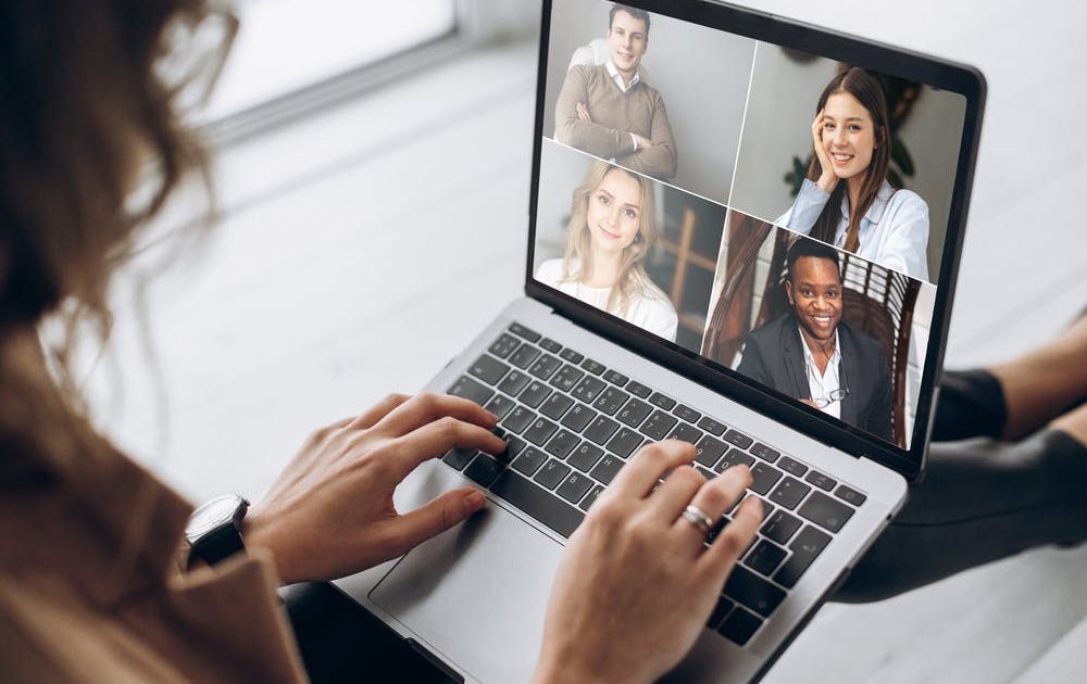 Een over-de-schouder weergave van een vrouw die deelneemt aan een videoconferentiegesprek op haar laptop, met vier glimlachende, diverse collega's op het scherm.