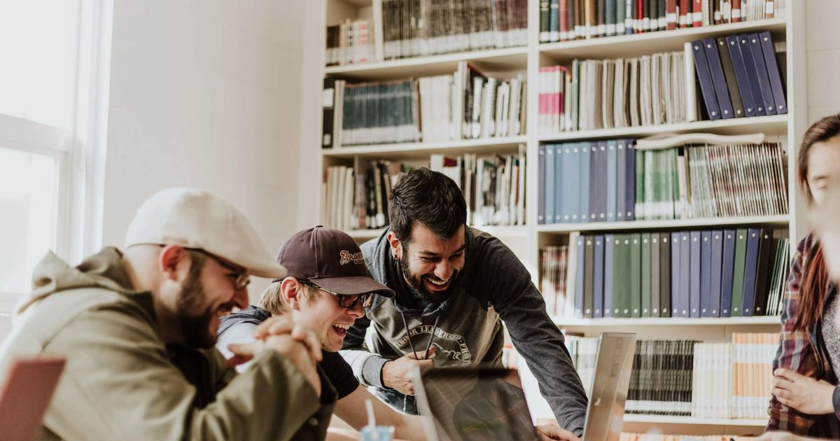 Een groep jonge studenten die samen lachen terwijl ze zich rond een laptop scharen om te studeren in een bibliotheek of klas vol boeken.