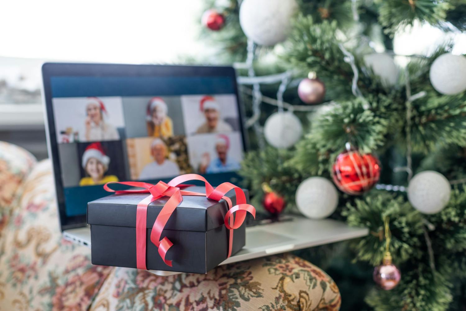 Une boîte cadeau noire ornée d'un ruban rouge posée sur un ordinateur portable, affichant un appel vidéo de groupe virtuel pour les fêtes, à côté d'un sapin de Noël décoré.