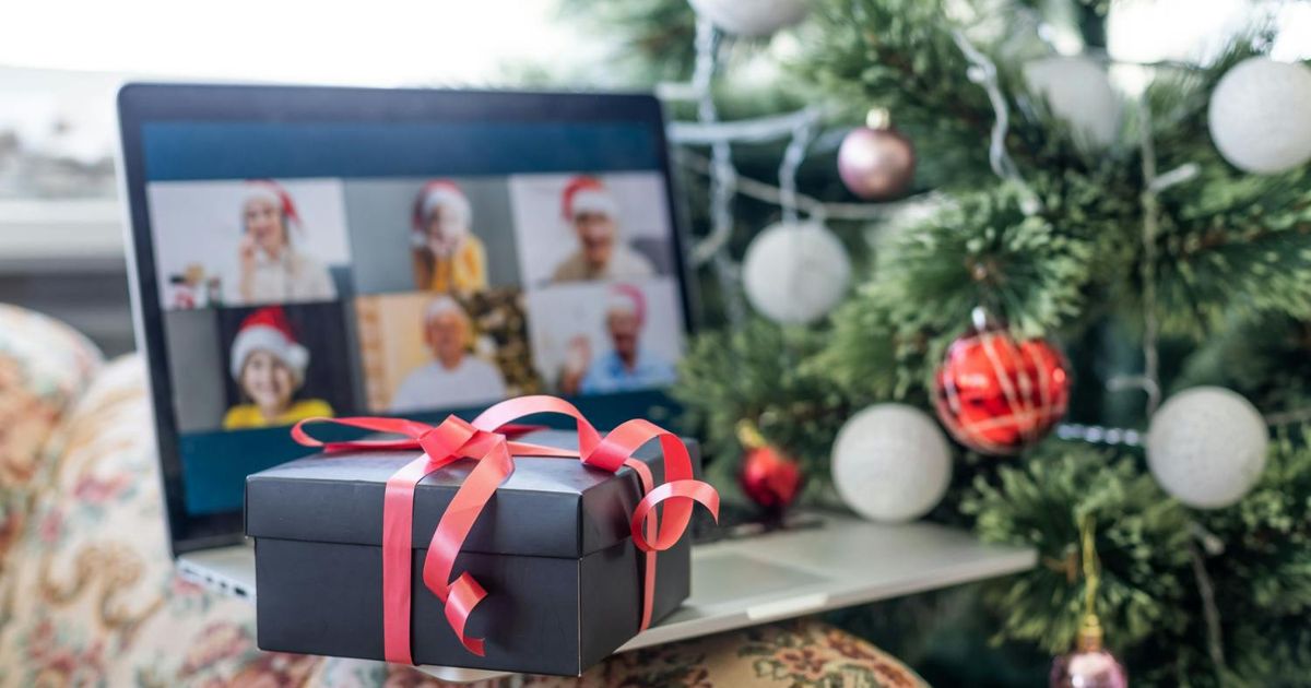 A black gift box wrapped with a red ribbon sitting on a laptop, which displays a virtual holiday group video call next to a decorated Christmas tree.