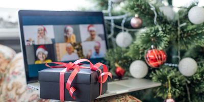 Een zwarte geschenkdoos met een rood lint op een laptop, waarop een virtuele feestelijke groepsvideogesprek wordt getoond naast een versierde kerstboom.