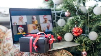 Een zwarte geschenkdoos met een rood lint op een laptop, waarop een virtuele feestelijke groepsvideogesprek wordt getoond naast een versierde kerstboom.