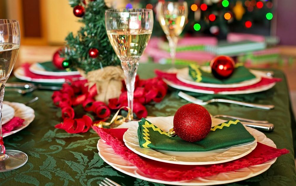 A Christmas dinner table laid for a group, with white scalloped plates on red and dark green napkins, a red glitter bauble resting on the nearest setting, filled champagne glasses, and curled red ribbon running down a green damask tablecloth in front of blurred Christmas tree lights.