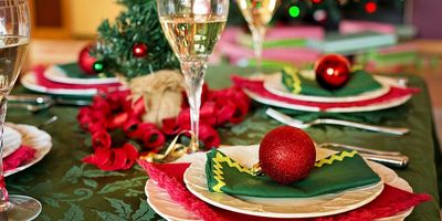 A Christmas dinner table laid for a group, with white scalloped plates on red and dark green napkins, a red glitter bauble resting on the nearest setting, filled champagne glasses, and curled red ribbon running down a green damask tablecloth in front of blurred Christmas tree lights.
