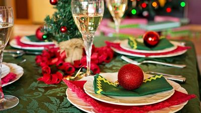 A Christmas dinner table laid for a group, with white scalloped plates on red and dark green napkins, a red glitter bauble resting on the nearest setting, filled champagne glasses, and curled red ribbon running down a green damask tablecloth in front of blurred Christmas tree lights.