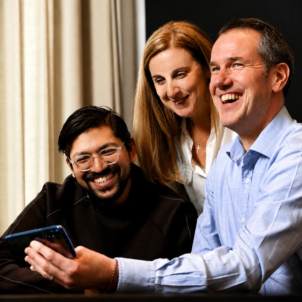 Een groep van drie lachende volwassenen die samen kijken naar een smartphone, wat duidt op een gedeelde, plezierige activiteit of spel.