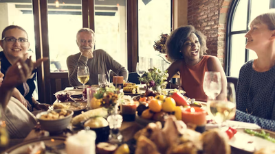A group of adults sit around a dining table filled with food, laughing and chatting together in a warm, cozy indoor setting with large windows and brick walls.