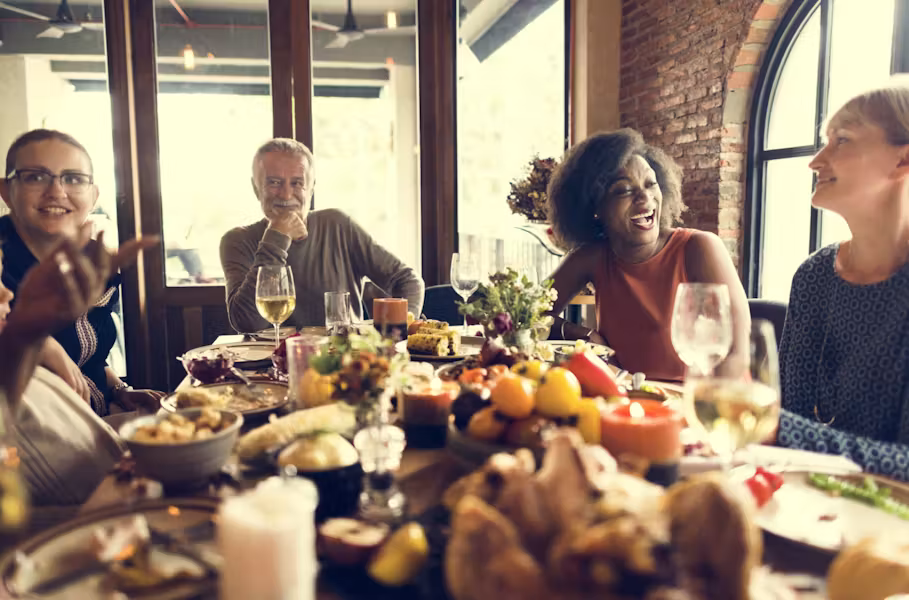 A group of adults sit around a dining table filled with food, laughing and chatting together in a warm, cozy indoor setting with large windows and brick walls.