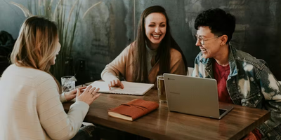 Three people sit around a table in a casual workspace, smiling and laughing while collaborating with a laptop and notebook, suggesting a friendly team meeting or brainstorming session.