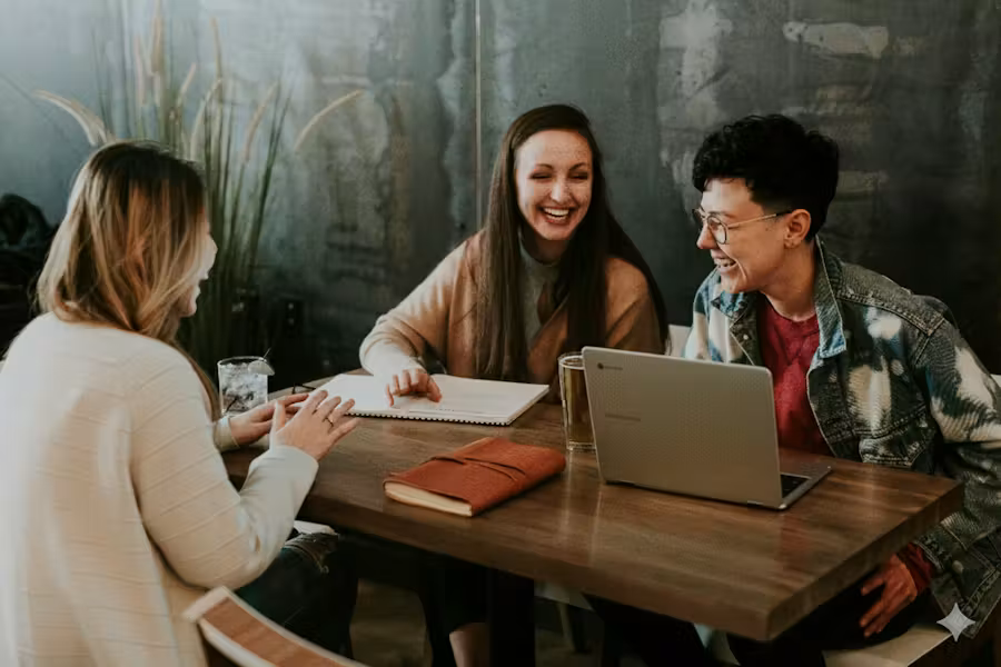Three people sit around a table in a casual workspace, smiling and laughing while collaborating with a laptop and notebook, suggesting a friendly team meeting or brainstorming session.