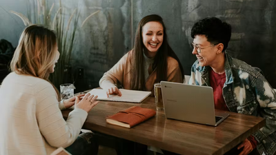 Three people sit around a table in a casual workspace, smiling and laughing while collaborating with a laptop and notebook, suggesting a friendly team meeting or brainstorming session.