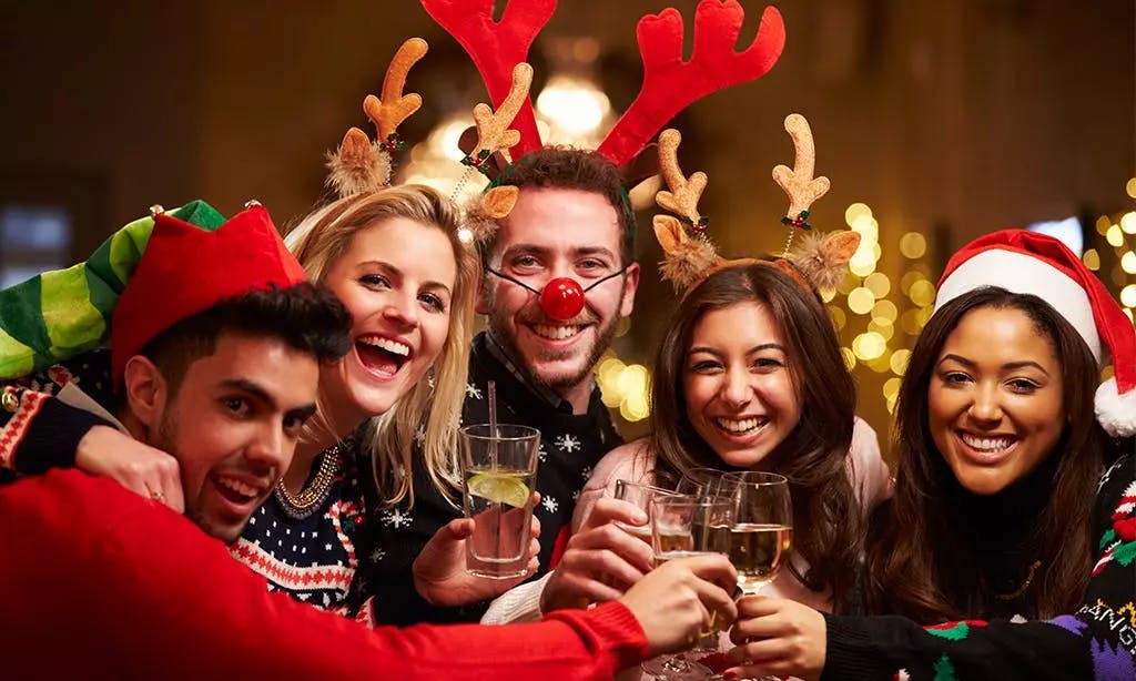 Un groupe joyeux de cinq amis souriant et trinquant ensemble lors d'une fête de Noël, portant des tenues festives comme des bois de renne, un bonnet de Père Noël, un chapeau de lutin et des pulls de Noël.