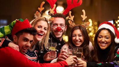 Een vrolijke groep van vijf vrienden die lachend klinken op een kerstfeest, gekleed in feestelijke outfits zoals rendiergewei, een kerstmuts, een elfenmuts en kersttrui.