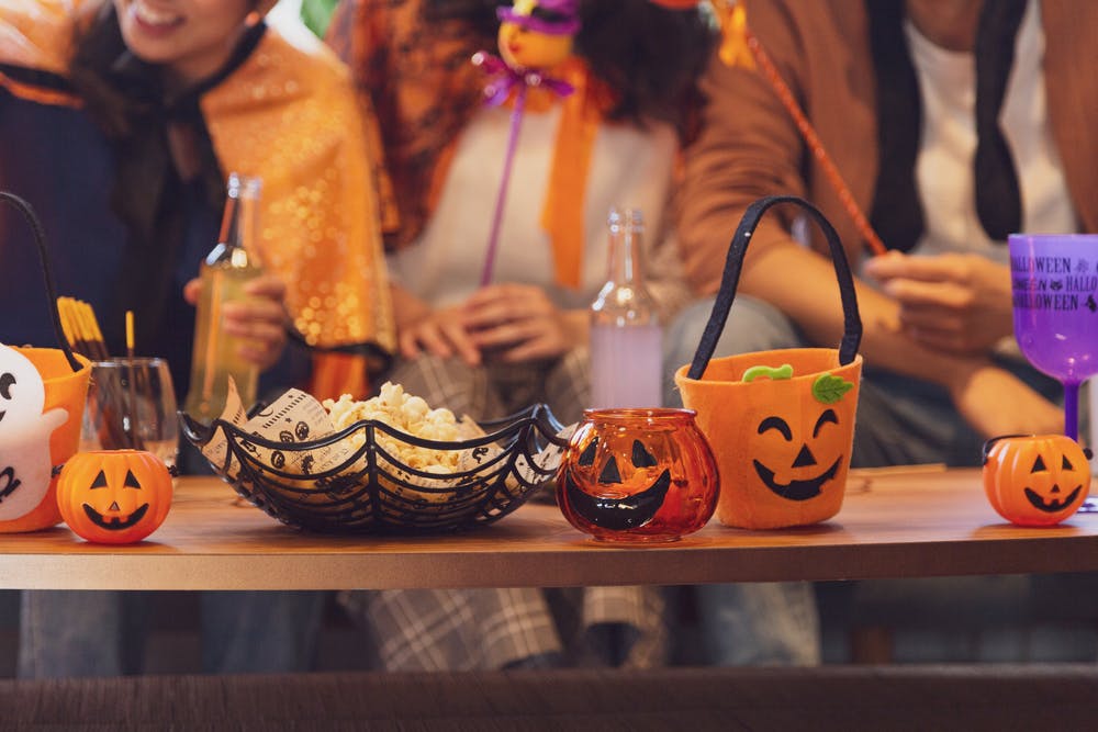 Gros plan d'une table en bois couverte de décorations d'Halloween, de popcorn dans un bol en toile d'araignée et de petits seaux citrouilles, avec des personnes en costume assises en arrière-plan flou.