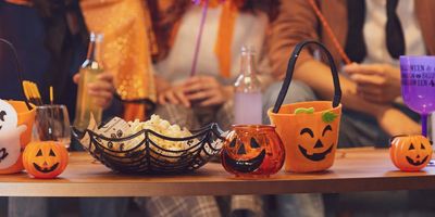 Een close-up van een houten tafel vol Halloween-decoraties, popcorn in een spinnenwebkom en kleine pompoenbakjes, met mensen in kostuums wazig op de achtergrond.