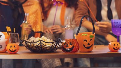 Een close-up van een houten tafel vol Halloween-decoraties, popcorn in een spinnenwebkom en kleine pompoenbakjes, met mensen in kostuums wazig op de achtergrond.