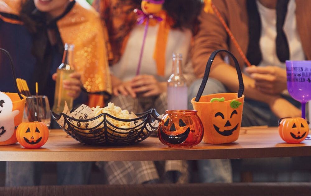 Een close-up van een houten tafel vol Halloween-decoraties, popcorn in een spinnenwebkom en kleine pompoenbakjes, met mensen in kostuums wazig op de achtergrond.