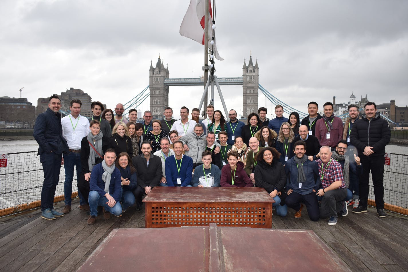 Un grand groupe de personnes souriantes et posant pour une photo sur le pont d'un bateau sur la Tamise, avec le célèbre Tower Bridge de Londres en arrière-plan sous un ciel nuageux.