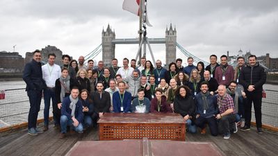 Een grote groep glimlachende mensen die poseren voor een foto op het dek van een boot op de Theems, met de beroemde Tower Bridge van Londen op de achtergrond onder een bewolkte hemel.