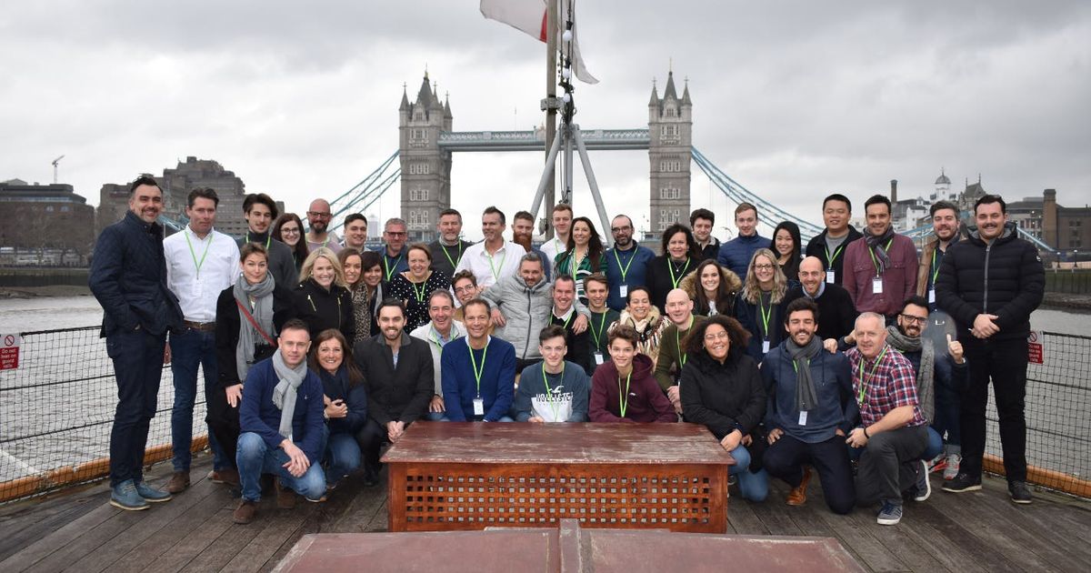 Een grote groep glimlachende mensen die poseren voor een foto op het dek van een boot op de Theems, met de beroemde Tower Bridge van Londen op de achtergrond onder een bewolkte hemel.