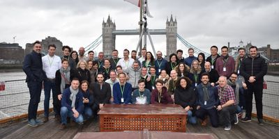 Een grote groep glimlachende mensen die poseren voor een foto op het dek van een boot op de Theems, met de beroemde Tower Bridge van Londen op de achtergrond onder een bewolkte hemel.