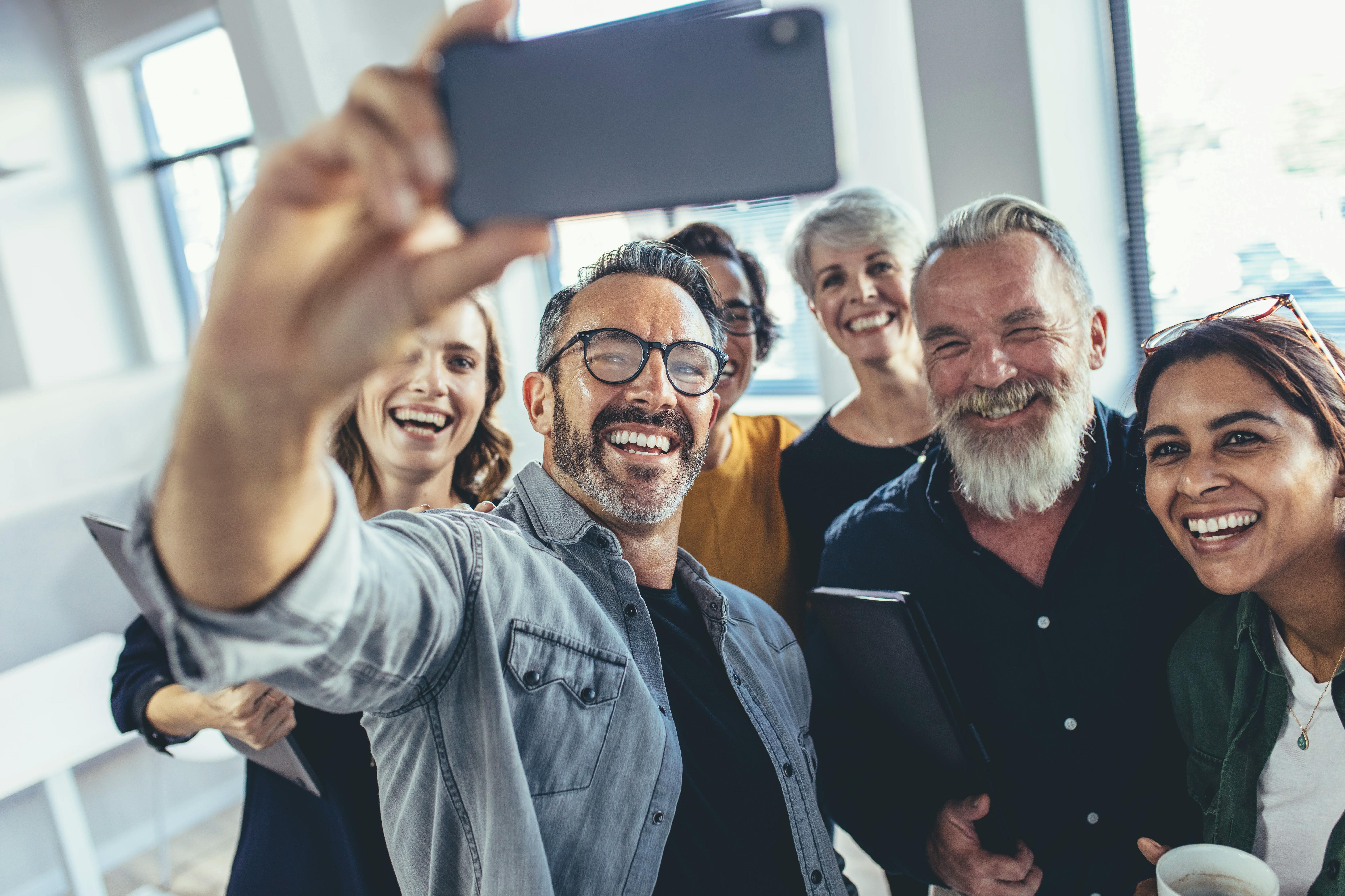 Un groupe souriant et diversifié de collègues adultes réunis dans un bureau lumineux, riant pendant qu'un homme lève un smartphone pour prendre un selfie de groupe.