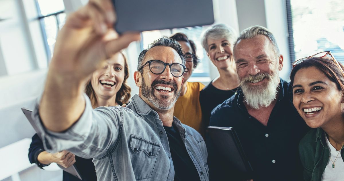 Een glimlachende, diverse groep volwassen collega's die samen in een licht kantoor bijeen zijn, lachend terwijl een man een smartphone omhooghoudt om een groepsselfie te maken.