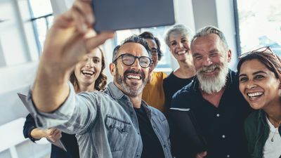 Een glimlachende, diverse groep volwassen collega's die samen in een licht kantoor bijeen zijn, lachend terwijl een man een smartphone omhooghoudt om een groepsselfie te maken.