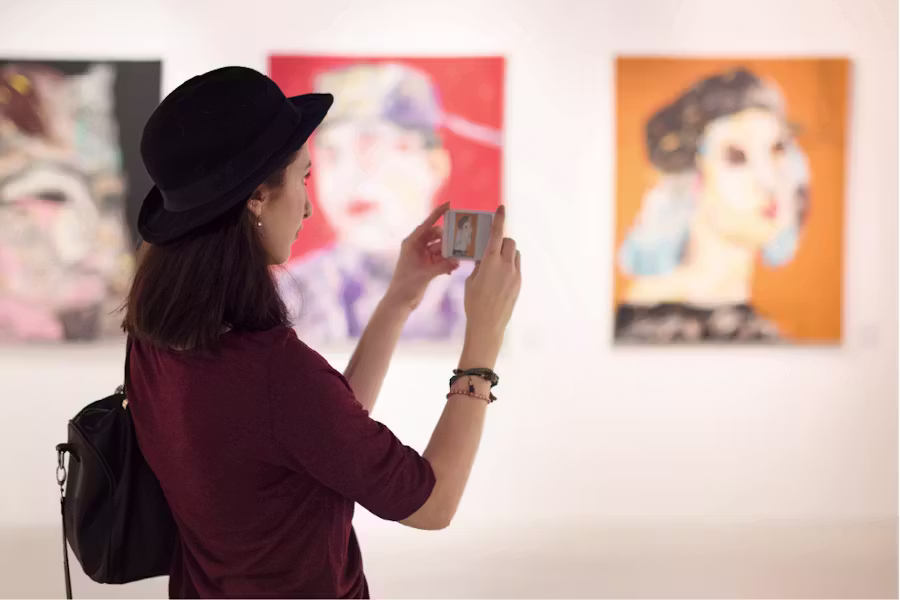 Een zijaanzicht van een vrouw met een zwarte bolhoed, die haar smartphone gebruikt om een foto te nemen van een kleurrijk schilderij in een kunstgalerie.