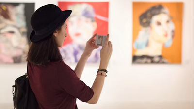 Een zijaanzicht van een vrouw met een zwarte bolhoed, die haar smartphone gebruikt om een foto te nemen van een kleurrijk schilderij in een kunstgalerie.