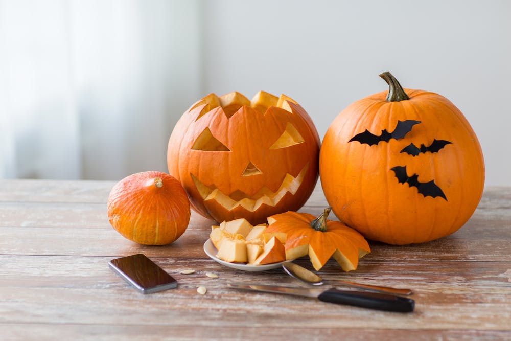Een close-up van een houten tafel vol Halloween-versieringen, popcorn in een spinnenweb-schaal en kleine pompoenemmers, met mensen in kostuums vaag op de achtergrond.